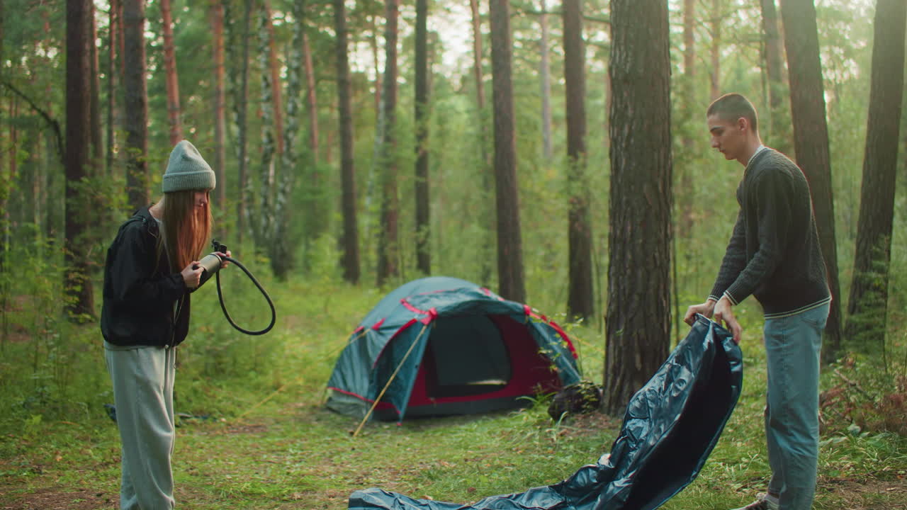 Young woman stands holding air pump in forest clearing while watching man stretch and shake tent fabric, surrounded by trees and greenery near pitched tent