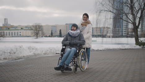 Fröhliche-Muslimische-Frau,-Die-Im-Winter-Mit-Ihrem-Behinderten-Freund-Im-Rollstuhl-Durch-Die-Stadt-Spaziert