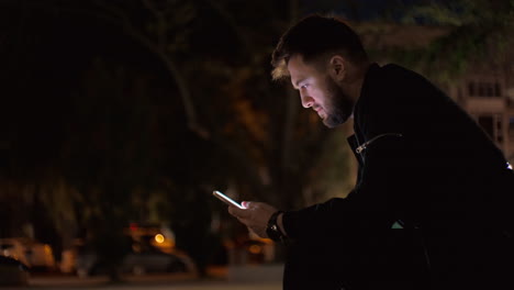 night time medium shot of a man sitting on a bench in a park, scrolling-searching on a smartphone