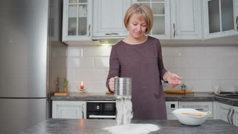 mujer tamiza la harina en la encimera mientras prepara la masa para hornear, el tamiz de acero inoxidable libera una nube suave de harina, el entorno de la cocina con iluminación cálida, utensilios de madera y cuenco de mezcla