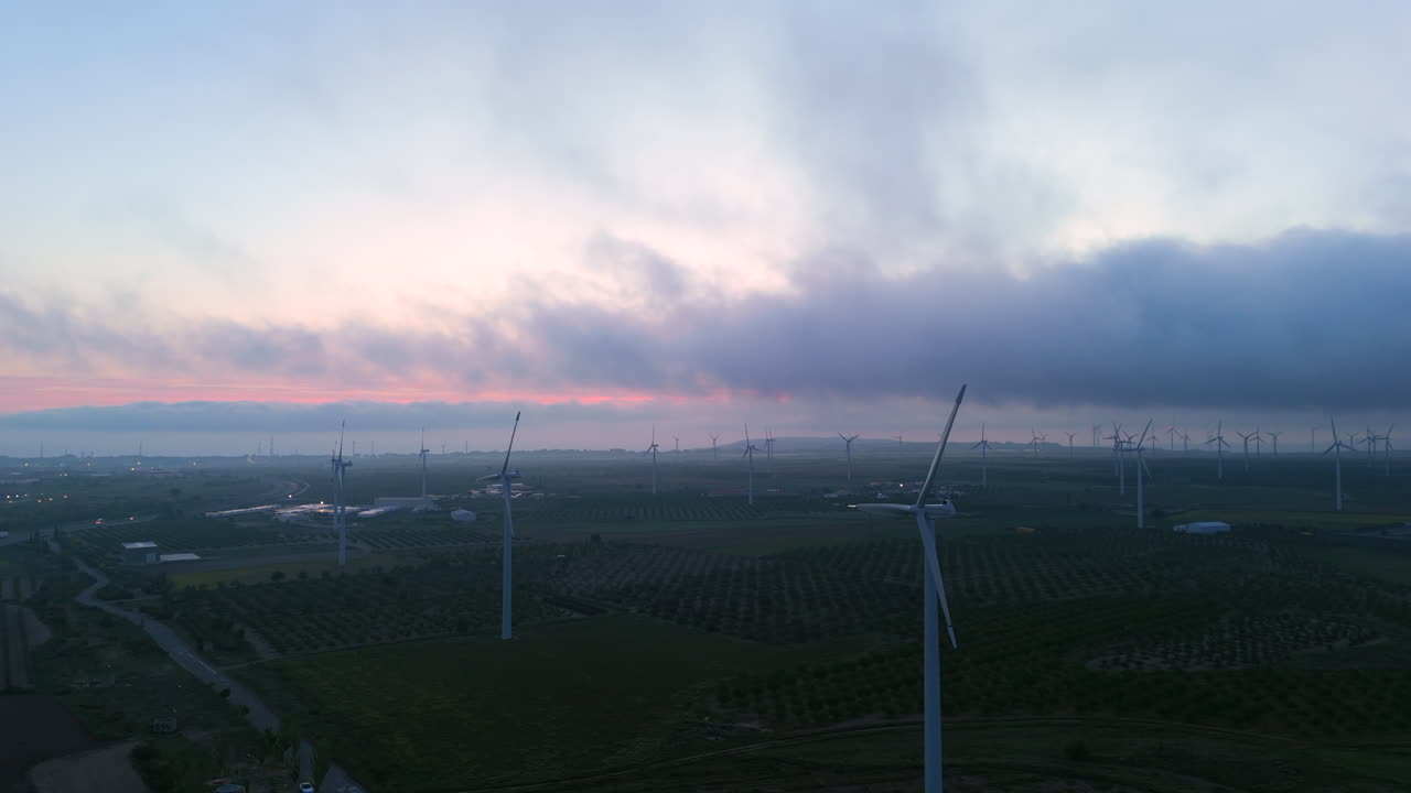 Aerial lateral ascent over a wind farm at dawn, showing dozens of wind turbines across cultivated fields and cloudy skies, captured with a drone in early morning light