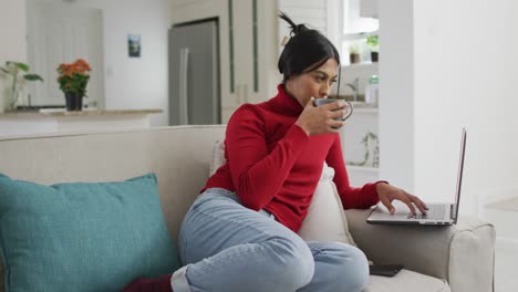 video de una feliz mujer biracial en hijab sentada en el sofá con café y usando una computadora portátil