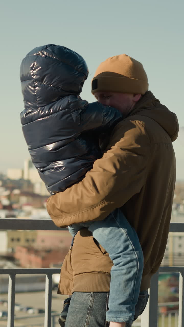 un padre sostiene tiernamente a su hijo cerca de una barandilla de hierro, ambos sonriendo mirándose cálidamente, con el paisaje de la ciudad a la vista, con ambos vestidos cálidamente con chaquetas
