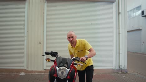 a bald man driving instructor in a yellow t-shirt drives a red moped into the garage