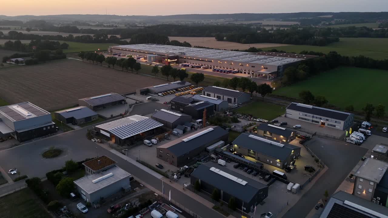 Industriegebäude mit Solarmodulen auf dem Dach bei Sonnenaufgang. Luftaufnahme im Überflug. Lagerhalle mit Lastwagen auf der Rampe. Felder in der Ferne. Weitwinkelaufnahme. Ruhiger Sonnenaufgang am Morgen.