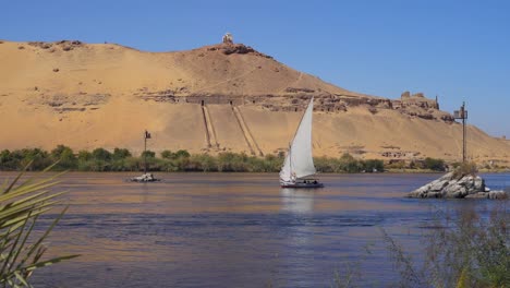 Sailboat-gliding-along-the-Nile-River,-showcasing-Aswan-Tombs-of-Nobles-and-serene-desert-landscapes-under-clear-blue-skies