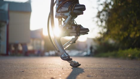 primer plano de las piernas en pilotes de muelle metálico saltando y girando al aire libre con zapatillas de deporte ligeras visibles. el fondo presenta una calle borrosa o un parque con árboles y coches en la suave luz del sol de la noche