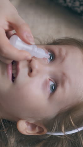 woman gives medical eye drops to suffering little girl lying on soft beige bed in light room extreme close upper view