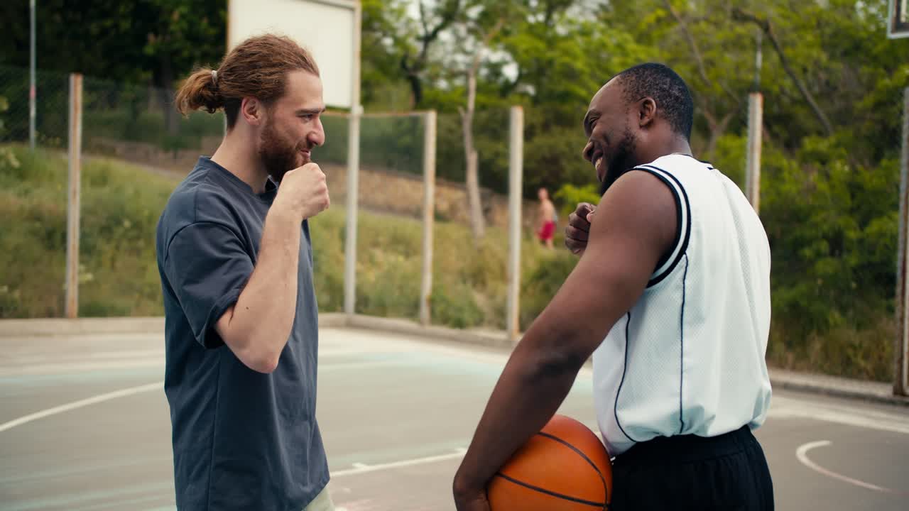 un hombre pelirrojo y su amigo, un hombre negro, se dan la mano y agitan sus puños de una manera amistosa en saludo. chicos duros en la cancha de baloncesto