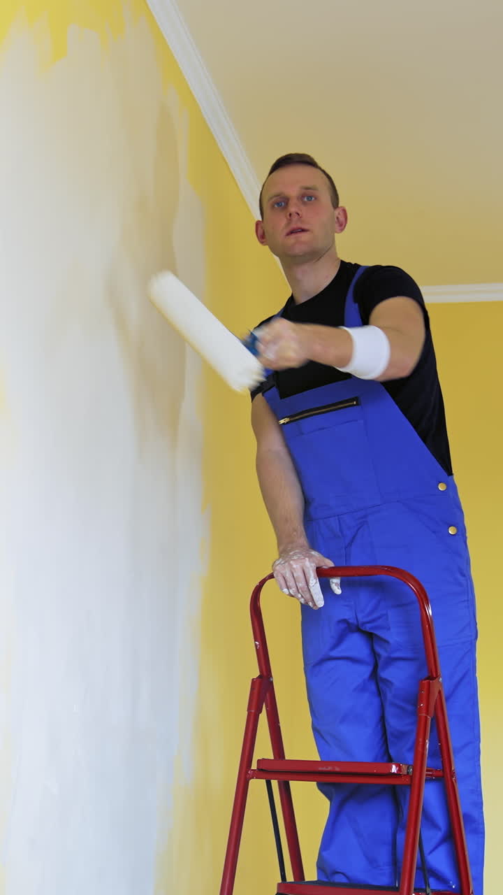 Concentrated male worker is painting room. Handsome man in work overalls stands on a ladder and painting wall with a roller white paint. Renovation in flat. Vertical video