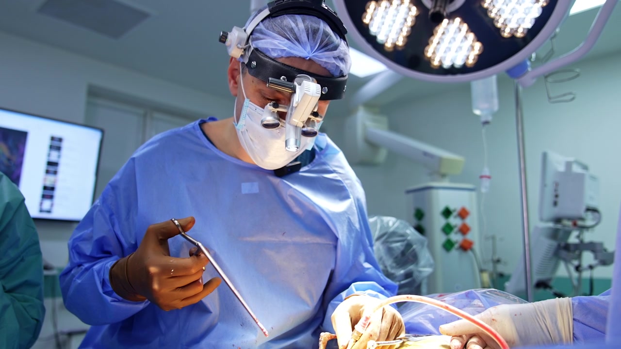Professionel mandlig neurokirurg holder et metalværktøj og et kirurgisk apparat. Læge med kirurgiske lupbriller udfører neurokirurgi