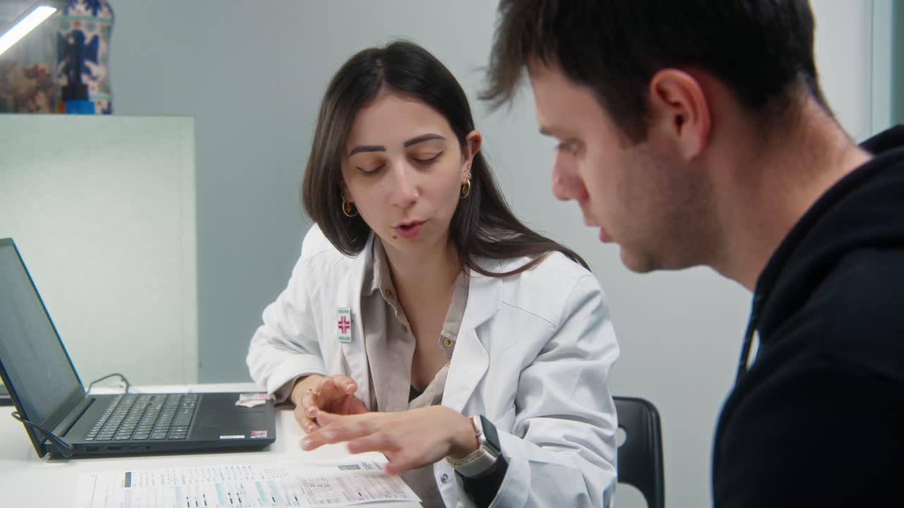 Bidikan jarak menengah seorang dokter wanita menjelaskan informasi medis kepada pasien pria selama konsultasi di kantor klinik modern.