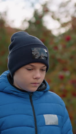 niño con gorra negra con parche y chaqueta azul paseando con la cabeza inclinada pensativamente, rodeado de follaje de otoño borroso y colores vibrantes