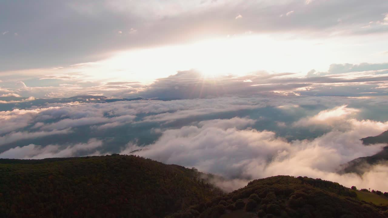Spettacolare vista aerea di un paesaggio nuvoloso in una valle sotto la montagna, con raggi solari che filtrano tra le nuvole e un sole bianco e brillante nel cielo blu