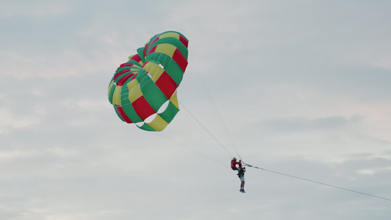 Parasailer in cerca di emozioni forti sopra la costa di un'isola, con una torsione dinamica della vela e cinghie dell'imbracatura tese, scia di motoscafo che si estende sotto, atmosfera da salto in alta quota, luce drammatica del crepuscolo e nuvole strutturate, energico.