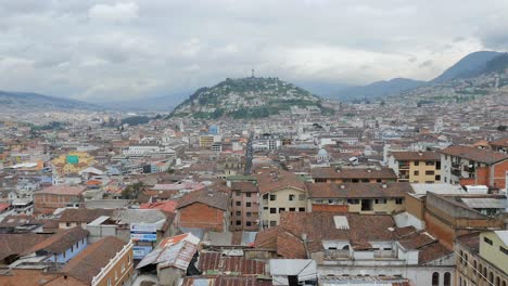 Spectacular-scenic-view-of-downtown-Quito-Equador-city-center,-buildings,-community-and-neighborhood-with-El-Panecillo-in-background-on-cloudy-sky-day,-handheld-pan-up