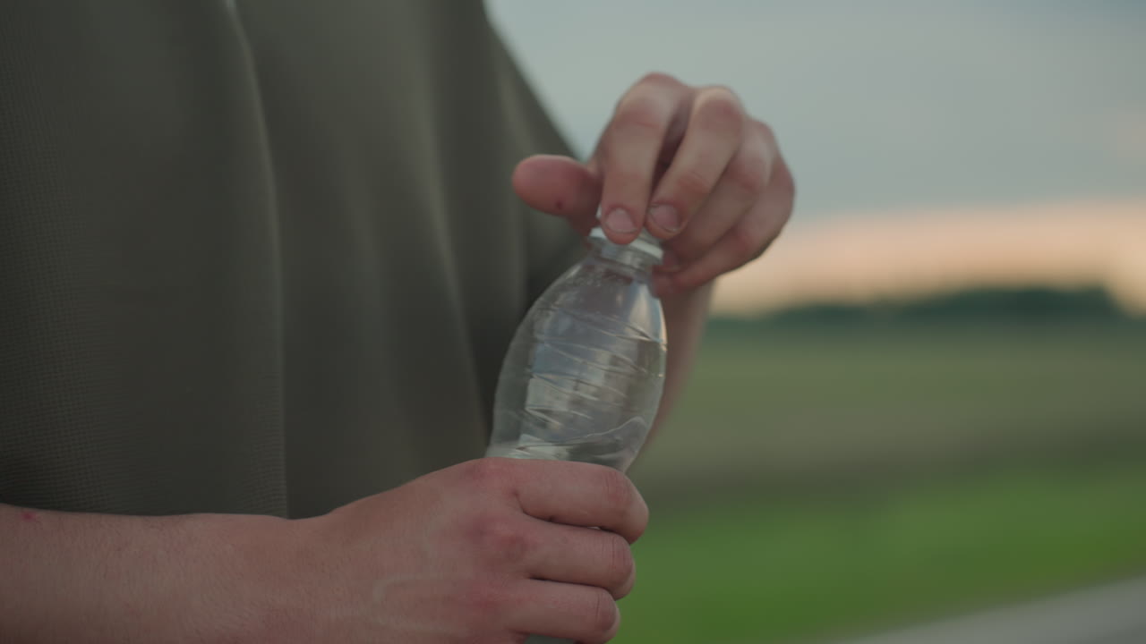 close up of hand twisting cap on clear water bottle against open road and field at sunset capturing plastic texture green shirt fabric and gentle wrist motion during outdoor hydration moment
