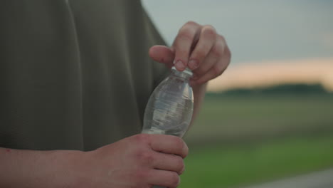 close up of hand twisting cap on clear water bottle against open road and field at sunset capturing plastic texture green shirt fabric and gentle wrist motion during outdoor hydration moment
