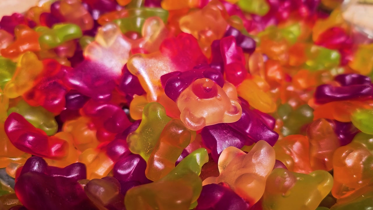 Pemandangan dekat tumpukan permen beruang gummy di toko permen, menunjukkan bentuk oranye, merah, kuning, ungu, dan hijau dengan permukaan mengkilap dan presentasi permen curah yang ceria