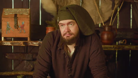 Disgusted barkeeper in cap stands in tavern. Emotional dwarf bartender looks at annoying guest in medieval store. Expressive bearded inn owner