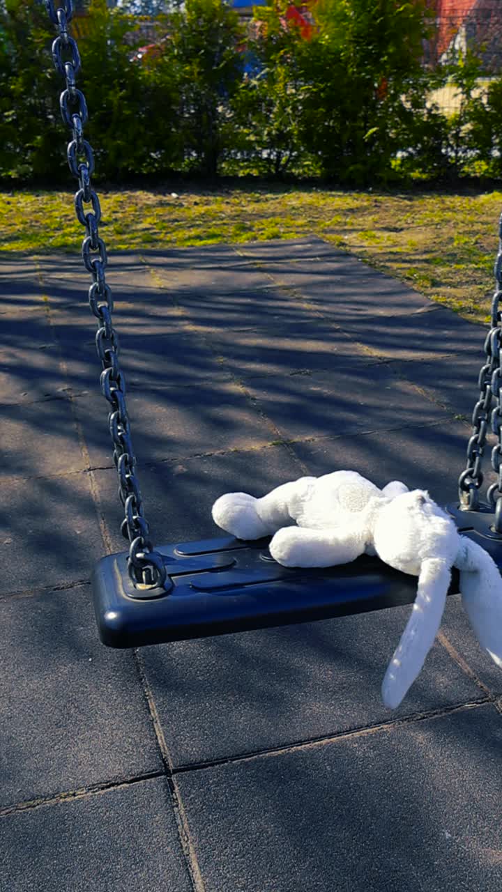 Coniglio di peluche bianco dimenticato che dondola da solo su un parco giochi vuoto, trasmettendo una sensazione di tristezza, solitudine, abbandono e ricordi d'infanzia perduti in una luminosa giornata di sole.