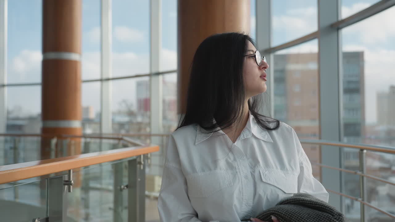 Young adult in white shirt walking through bright modern mall interior holding gray checkered coat, calmly looking around as sunlight streams through tall glass panels showcasing urban cityscape