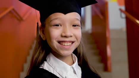 Zoom-In-Of-A-Happy-Preschool-Female-Student-In-Cap-And-Gown-Holding-Graduation-Diploma-And-Looking-At-The-Camera-1