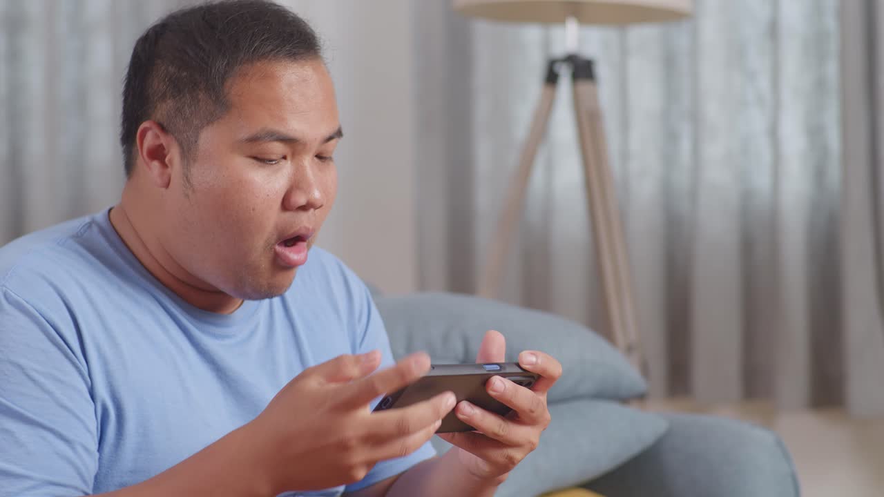 한 아시아 남자가 케과 함께 김을 먹고 집에서 거실 소파에서 패스트푸드를 먹는 동안 스마트폰에서 게임을 하고 있습니다.