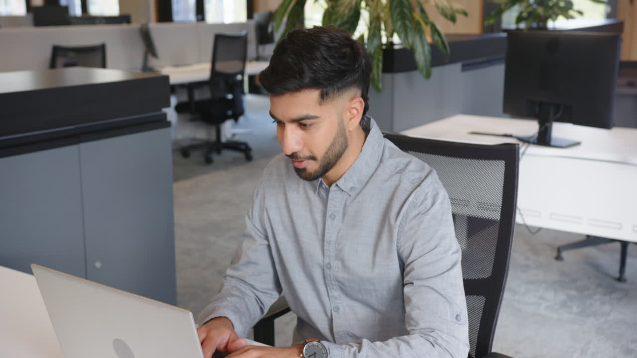 Working in modern office, Indian man using laptop and smiling at desk