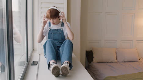 Girl-Sitting-On-Windowsill-In-The-Bedroom,-Then-She-Puts-The-Headphones-And-Plays-Music-From-Her-Smartphone