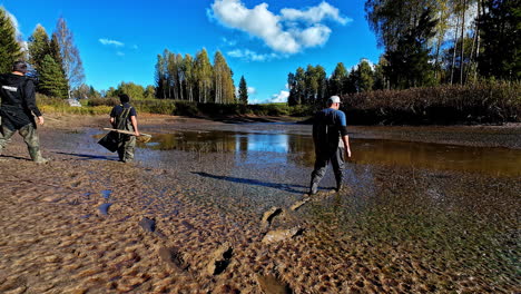 men in waterproof fishing jumpsuit walking on a muddy and drained fish pond to catch fish. - wide shot
