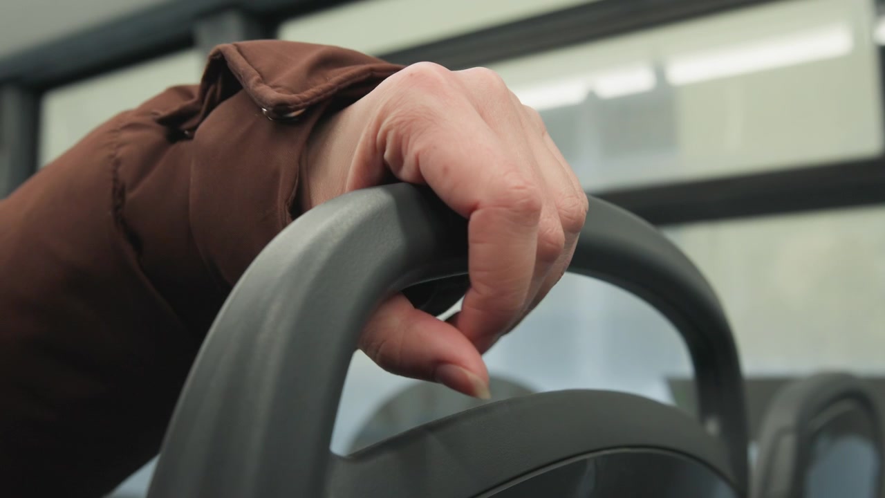 Primer plano de la mano de un pasajero agarrando firmemente el asa superior del reposacabezas del asiento dentro de la cabina de un autobús o tren, con tensión en los dedos y un agarre firme que sugiere apoyo, equilibrio, comodidad y disposición para ponerse de pie