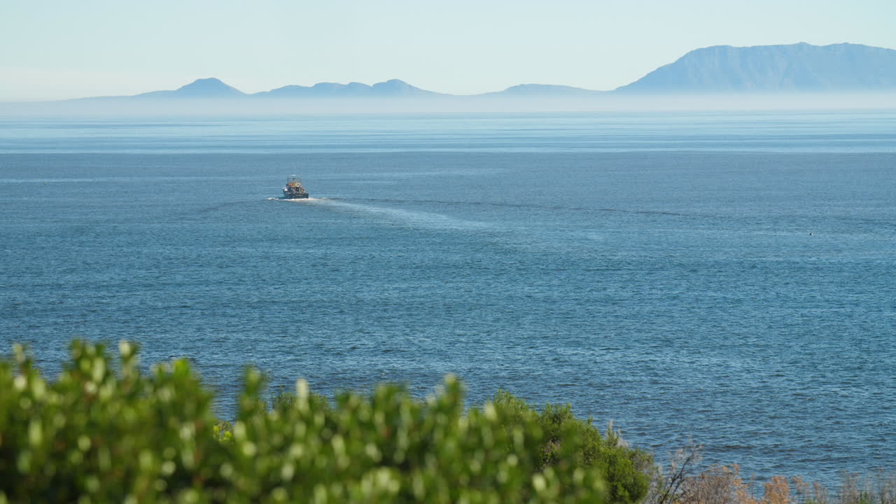 Fishing trawler sailing away in blue waters off the coast of South Africa on clear sunny day with heatwaves and green shrubbery in foreground and misty mountains in background