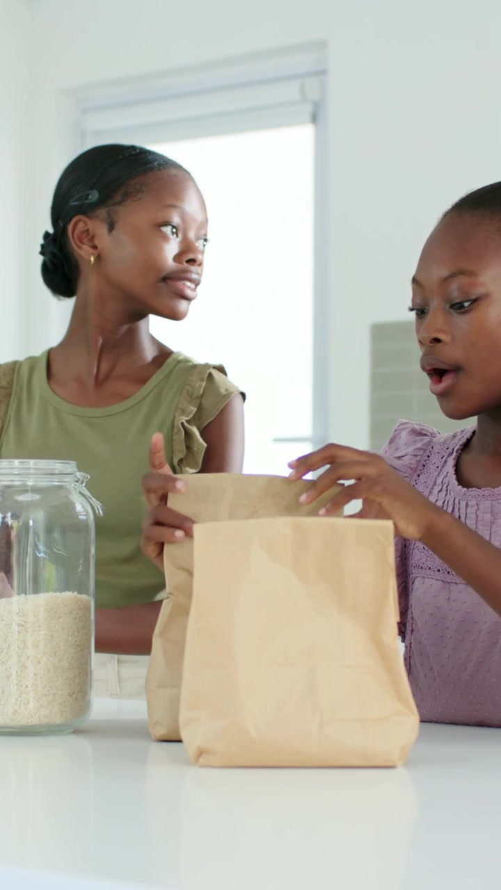 Vertical video: Reaching bag, duo in purple green filling brown bags at counter to pack with jar