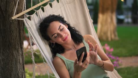 Portraitof-A-Woman-Relaxing-In-A-Hammock-Outdoors,-Surfing-Internet-In-Her-Smartphone,-Clicks-On-The-Screen-And-Smiling