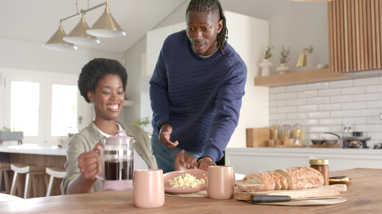 Pouring coffee into mugs, african american woman smiling while man stands nearby in kitchen