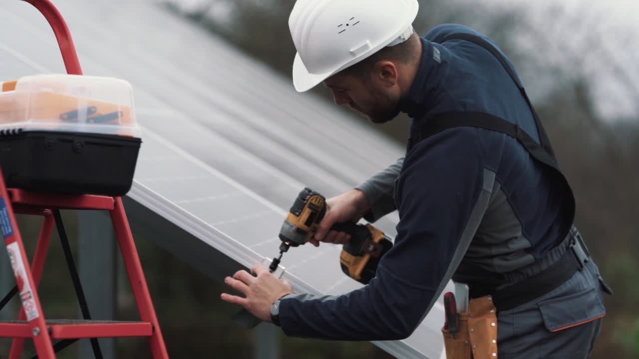 Close-up of a young man in a work uniform installing a solar panel with a cordless screwdriver