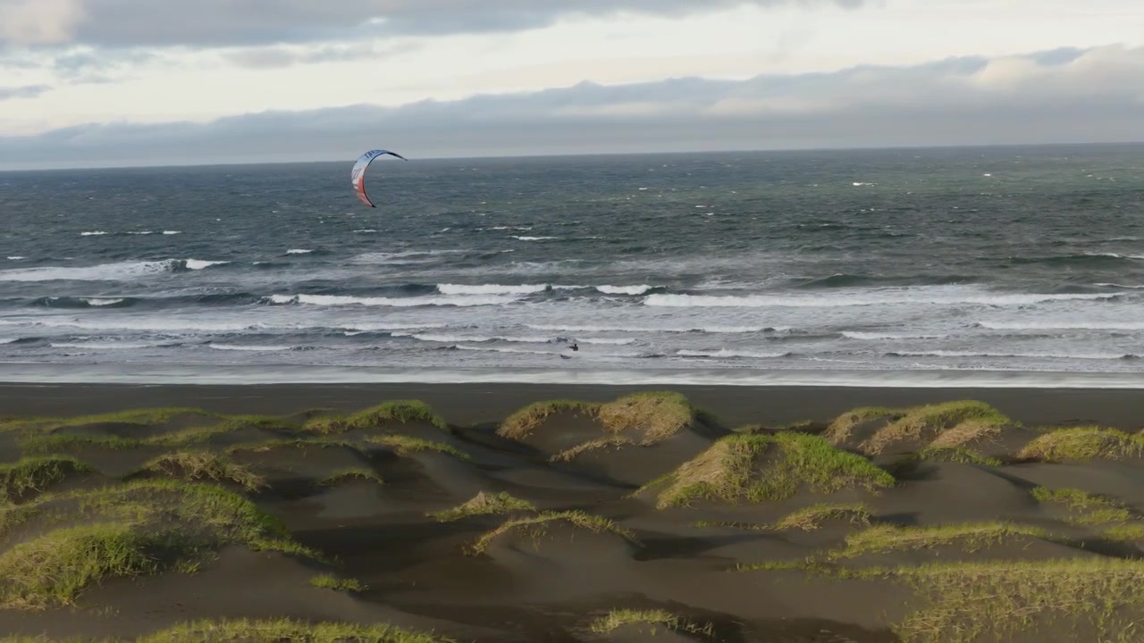 Kitesurfista surfa ondas do Atlântico impulsionadas pelo vento além de dunas de areia vulcânica preta e vegetação costeira na costa sul da Islândia sob montanhas distantes, rastreamento por drone