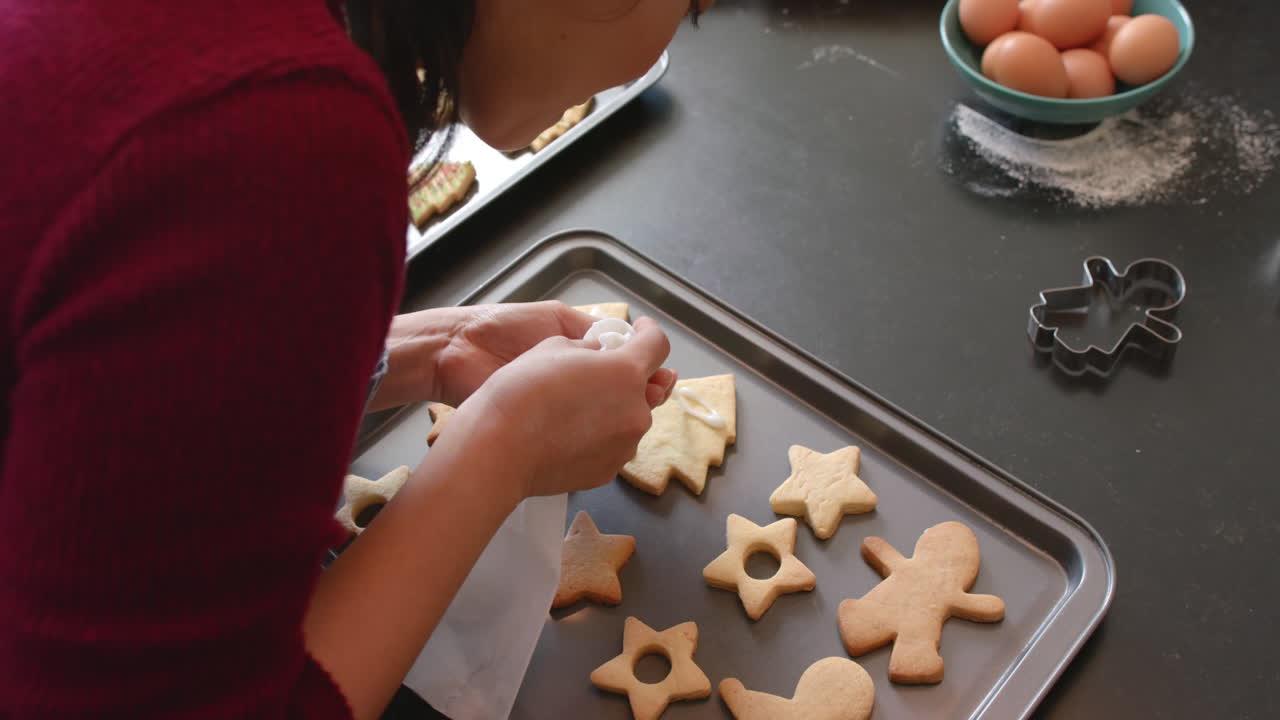 feliz mujer biracial en delantal decorando galletas de navidad en la cocina, cámara lenta
