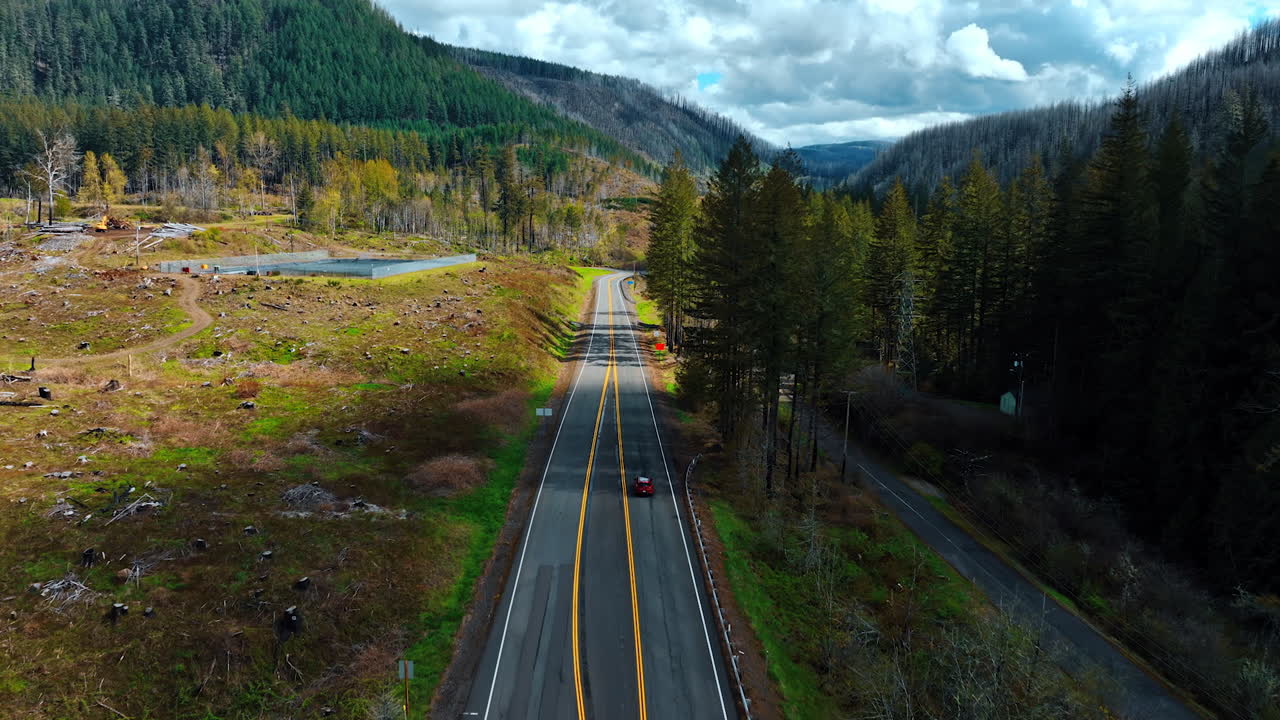 Following the red automobile riding by the highway along the beautiful nature. Mountains overgrown with pine trees at backdrop. Top view.