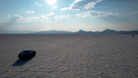 Drone-overtake-car-riding-across-Bonneville-Salt-Flats-unique-natural-features-with-beam-of-sunlight-rays-shining-through-dramatic-clouds-and-mountain-touching-horizon-in-background,-Utah