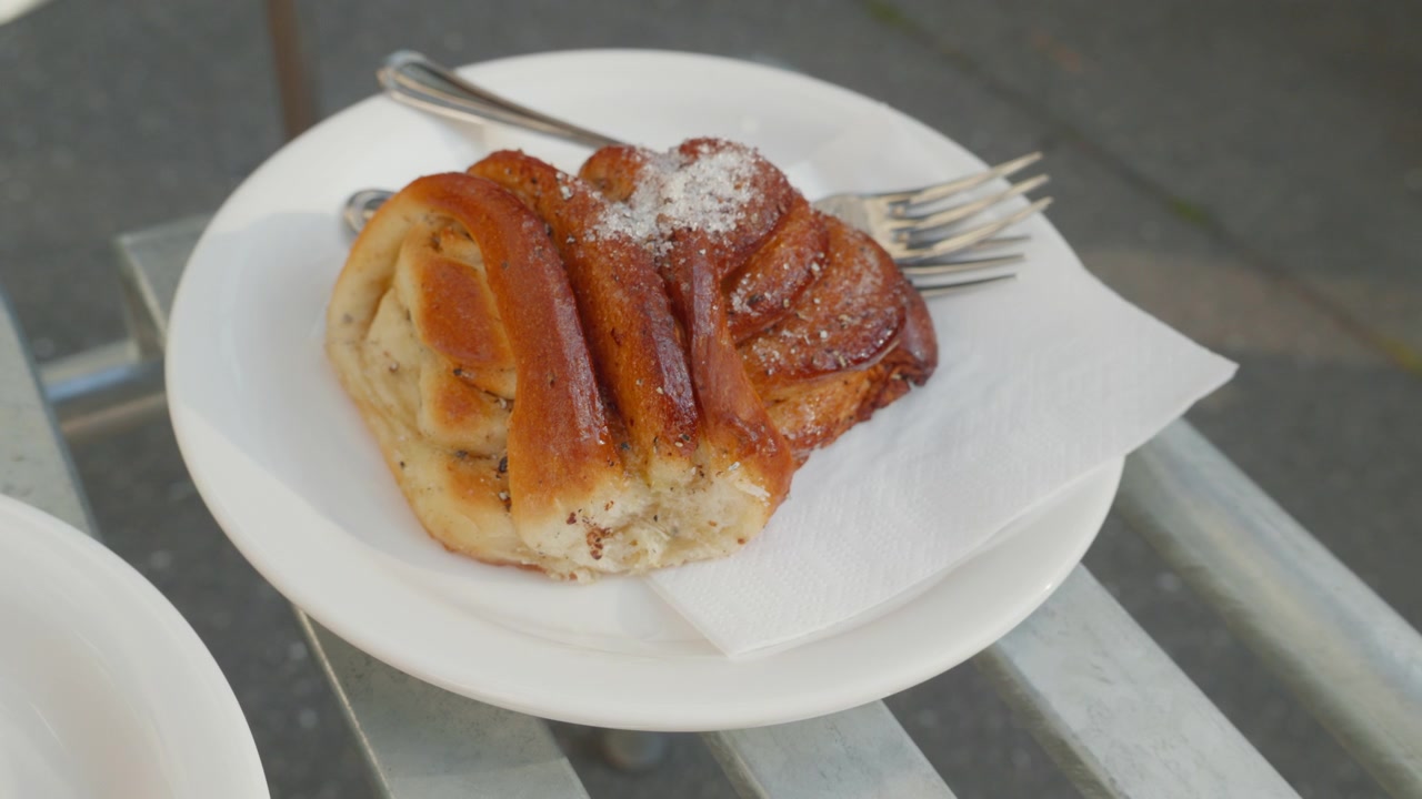 Un dolce panino al cardamomo servito su un piatto bianco all'aperto su una panchina stradale assolata, perfetto per temi di panetteria, pausa caffè e cultura culinaria urbana
