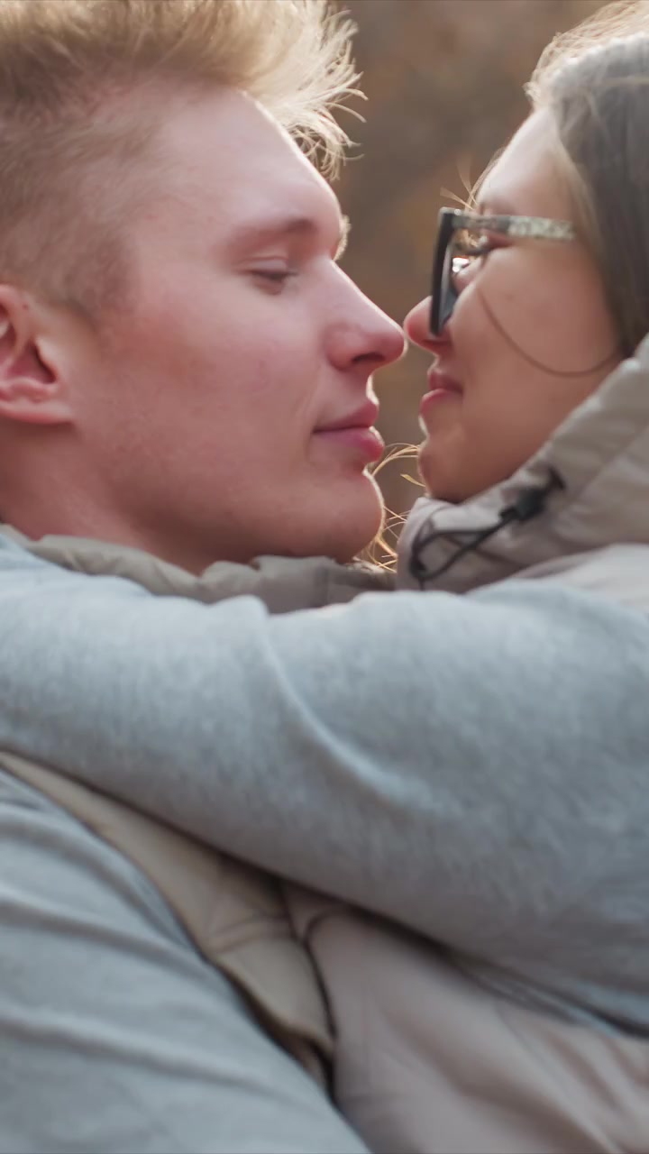 Woman with long brown hair wearing glasses and beige vest. Interaction includes gentle touch between two subjects. Wind causes slight movement of hair on both individuals