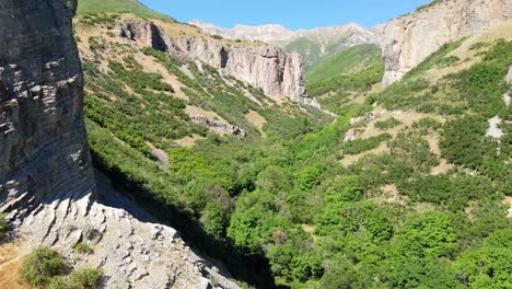 Flying-close-to-a-shear-rugged-cliff-with-a-view-of-a-canyon-and-vast-mountain-range