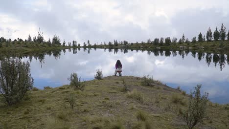 a woman seated near lakeshore with mirror reflection in huaraz, peru. static shot