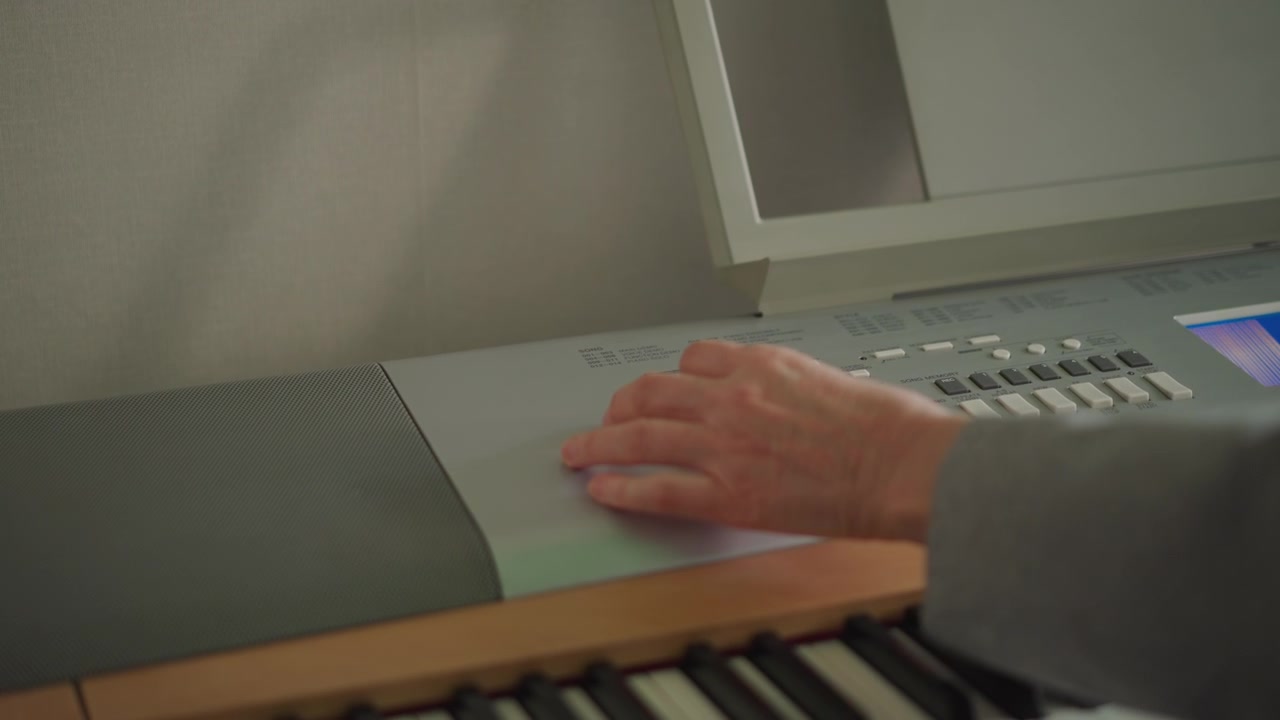 Insegnante di tastiera che regola il controllo del sintetizzatore in studio, inquadratura sfocata da dietro la spalla che mostra la mano sul pannello di controllo e sulla tastiera, momento di insegnamento calmo con luce ambientale calda, display elettronico discreto.