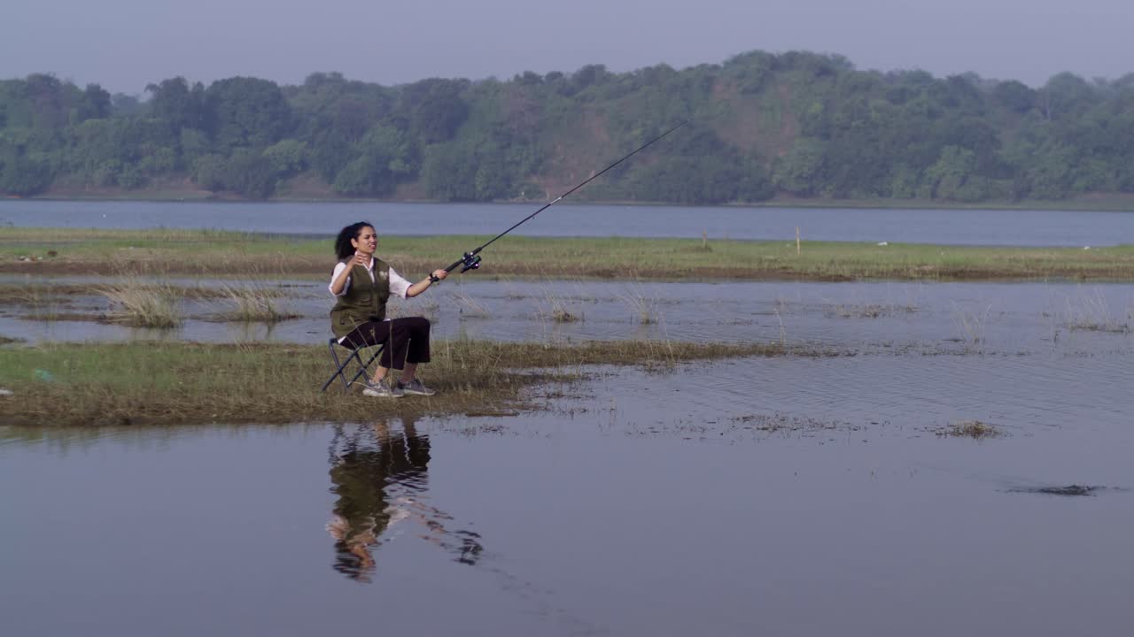 Seorang gadis Asia Selatan mengenakan rompi luar ruangan, duduk, fokus menyesuaikan gulungan pancingnya selama momen tenang di tepi danau.