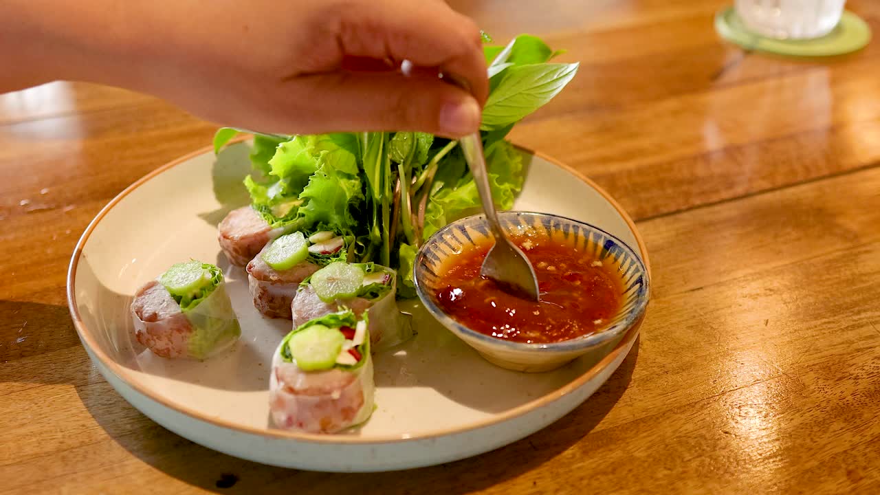A hand mixes sauce beside Vietnamese pork rolls on a wooden table, highlighting culinary preparation in a Bangkok restaurant