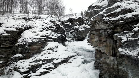 Drone-flying-between-rocky-frozen-river-banks-with-bridge-in-background,-Norway
