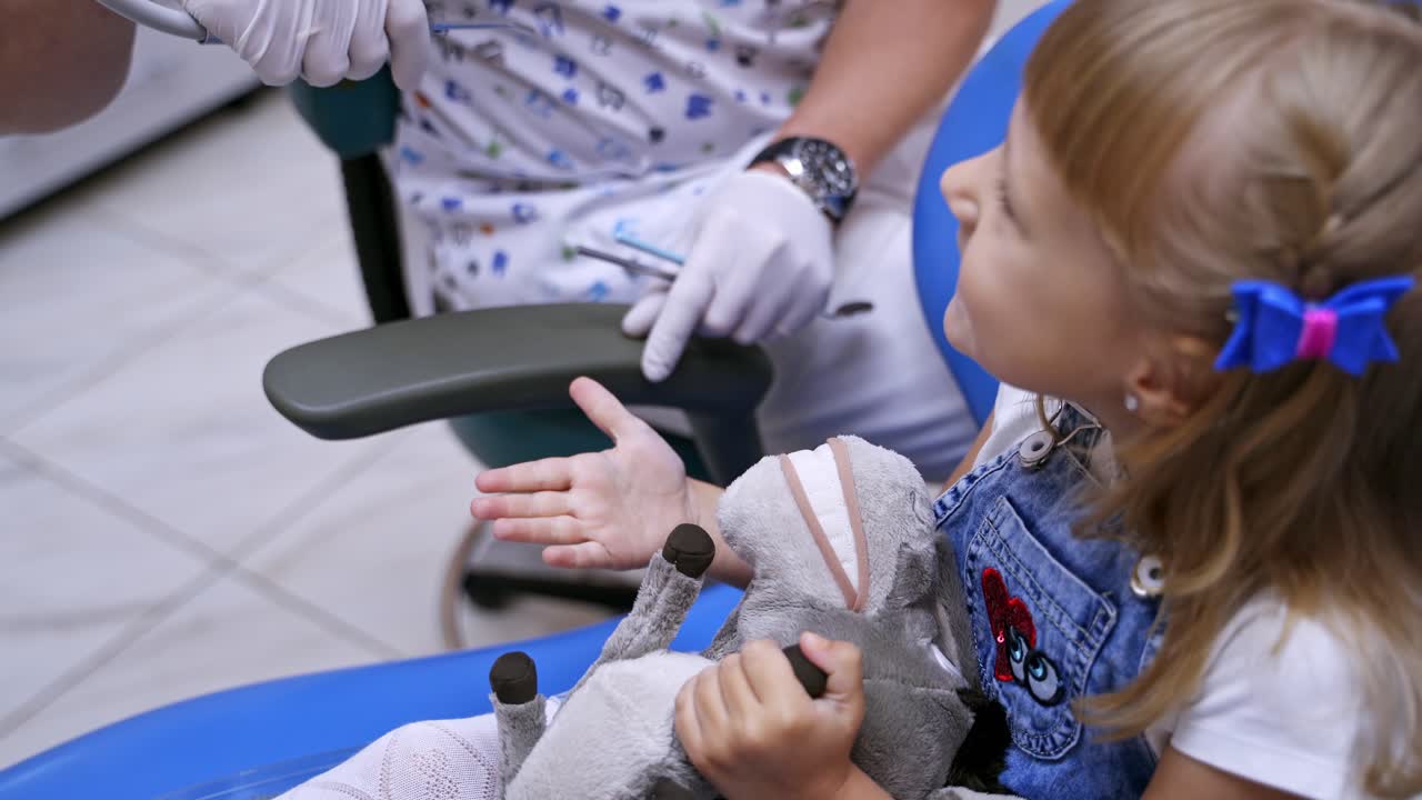 Little child in stomatology chair - close up. Cute girl with a toy at dentist cabinet. Courageous baby explaining orthodont her problem. Closeup video.